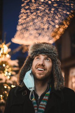 Kürk şapkalı ve yüz maskeli çekici bir adam Noel kutlamaları sırasında ve 2021 yılbaşı arifesinde Coronavirus covid-19 pandemisi sırasında aydınlatılmış şehir ışıkları altında.