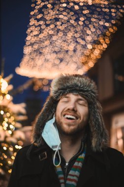 Kürk şapkalı ve yüz maskeli çekici bir adam Noel kutlamaları sırasında ve 2021 yılbaşı arifesinde Coronavirus covid-19 pandemisi sırasında aydınlatılmış şehir ışıkları altında.