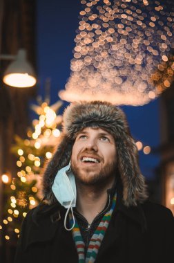 Kürk şapkalı ve yüz maskeli çekici bir adam Noel kutlamaları sırasında ve 2021 yılbaşı arifesinde Coronavirus covid-19 pandemisi sırasında aydınlatılmış şehir ışıkları altında.