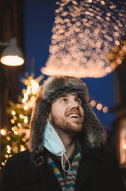 Kürk şapkalı ve yüz maskeli çekici bir adam Noel kutlamaları sırasında ve 2021 yılbaşı arifesinde Coronavirus covid-19 pandemisi sırasında aydınlatılmış şehir ışıkları altında.