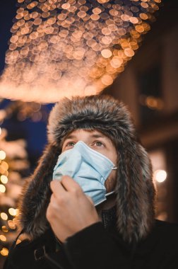 Kürk şapkalı ve yüz maskeli çekici bir adam Noel kutlamaları sırasında ve 2021 yılbaşı arifesinde Coronavirus covid-19 pandemisi sırasında aydınlatılmış şehir ışıkları altında.