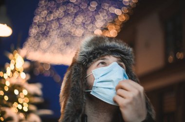 Kürk şapkalı ve yüz maskeli çekici bir adam Noel kutlamaları sırasında ve 2021 yılbaşı arifesinde Coronavirus covid-19 pandemisi sırasında aydınlatılmış şehir ışıkları altında.