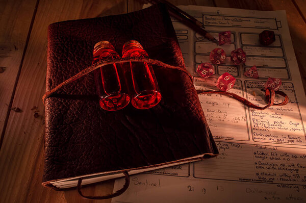 Image of a leather bound tome with on top two bottles with red liquid. Role playing dice, pencils and a note sheet for table top gaming