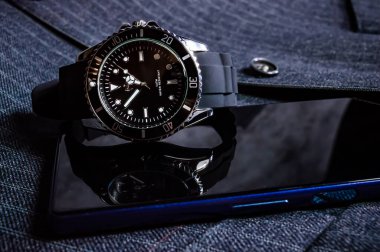 Siyah krom bir erkek saati ve yelek giymiş mavi bir telefon yakın plan.