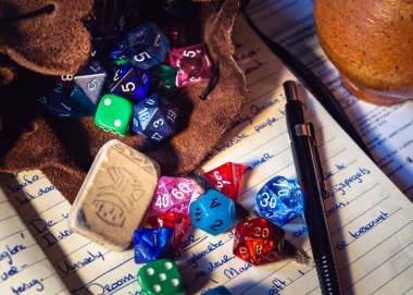 Karakter kağıdındaki deri bir zar çantasından dökülen çeşitli şekil ve renklerdeki zar oyunlarının yakın plan görüntüsü. Arka planda seramik bir içecek kavanozu, bir kalem ve silgi var.