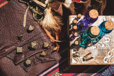 Ahşap bir kutunun üzerinde renkli sıvı bulunan cam durdurucu şişelerin, deri kaplı bir kitabın üzerinde metalik RPG oyun zarları ve güneşteki deri bir zar çantasının bir parçası.