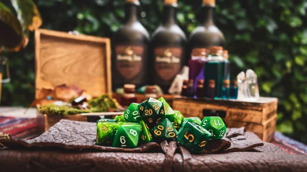 Deri kaplı bir kitabın üzerinde yeşil RPG oyun zarları yığını. Arka plandaki ahşap kutuların içinde çeşitli renklerde sıvılar var.