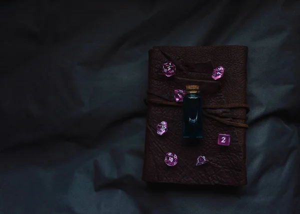 Image of pink transparent gaming dice and a glass vial with dark liquid on a leather-bound notebook in the shadow