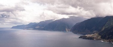 Sisli dağ kıyıları, bulutlu tepeler huzurlu deniz manzarasını sarar, Puslu dağ kenarı heybetli bir şekilde sakin su manzarasını sarar, sisli zirveler ve sessiz kayalıklar sakin ve karamsar sahil manzarası yaratır