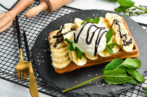 Belgian waffles: with banana, ice cream and chocolate. Dessert. On a black stone plate.