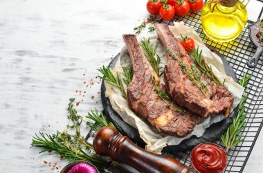 Kemiğin üzerinde ızgara dana biftek, beyaz ahşap arka planda baharatlar ve sebzeler. Düz yatıyordu. Üst görünüm.