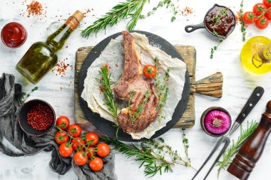 Kemiğin üzerinde ızgara biftek, beyaz ahşap arka planda baharatlar ve sebzeler. Düz yatıyordu. Üst görünüm.