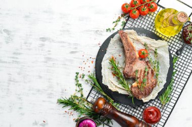Kemiğin üzerinde ızgara biftek, beyaz ahşap arka planda baharatlar ve sebzeler. Düz yatıyordu. Üst görünüm.