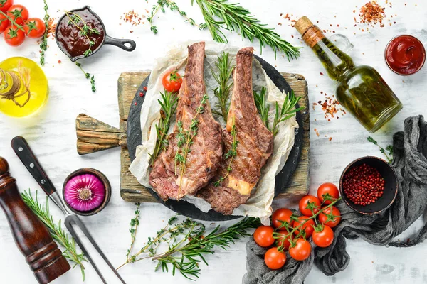 Kemiğin üzerinde ızgara dana biftek, beyaz ahşap arka planda baharatlar ve sebzeler. Düz yatıyordu. Üst görünüm.