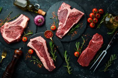 Çiğ biftek seti - T-bone, tomahawk, striploin, fileto, New York bifteği. Siyah taştan bir arka planda. Üst görünüm.
