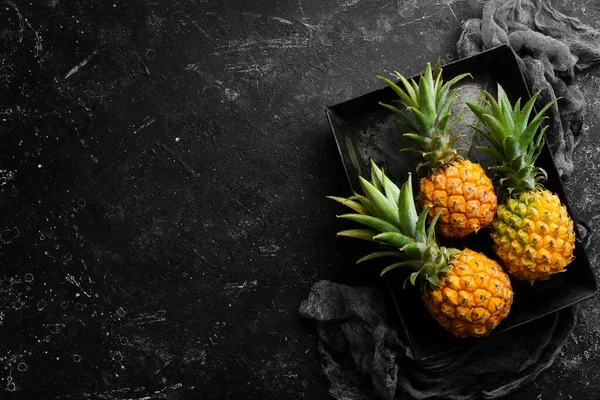 Pineapples in a metal box on a black stone background. Ripe baby pineapple. Tropical fruits. Top view. Free space for text.
