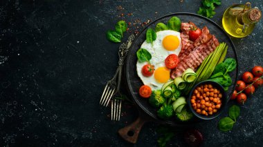 İngiliz kahvaltısı: nohut, avokado, çırpılmış yumurta, siyah taş tabakta domuz pastırması sebzesi. Üst Manzara. Metin için boş alan.