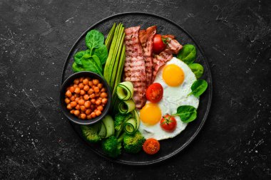 İngiliz kahvaltısı: nohut, avokado, çırpılmış yumurta, siyah taş tabakta domuz pastırması sebzesi. Üst Manzara. Metin için boş alan.