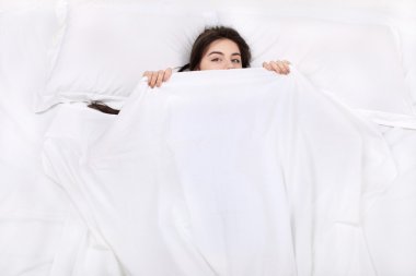 Top view photo of beautiful mixed-race girl lying on big white bed. Young pretty woman wearing pajamas. Girl lying on back, looking at camera and covering herself with blanket clipart