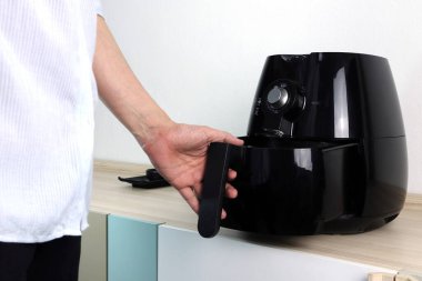 Sol el, aile üyelerine yemek pişirmek için beyaz beton duvar arka planına sahip beyaz mutfaktaki ahşap masanın üzerinde duran siyah hava fritözünü ya da yağsız fritöz aletini tutuyor.