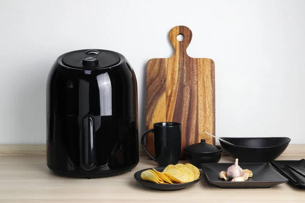 a black deep fryer or oil free fryer appliance, mug, dish and wooden tray are on the wooden table in the kitchen with a small plant in the pot ( air fryer )