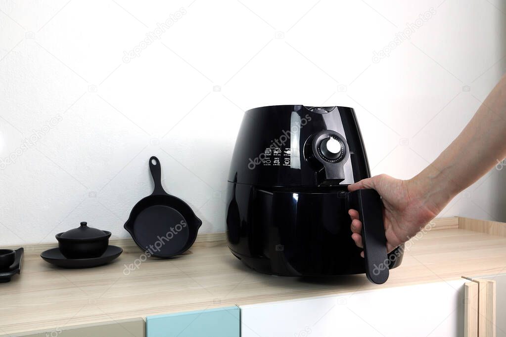 the right hand is holding the black air fryer or oil free fryer appliance which is on the wooden table in the white kitchen with white cement wall background for cooking the dinner for family members