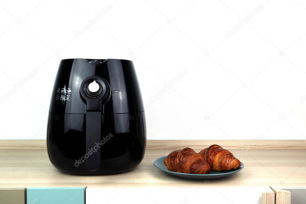 the black air fryer or oil free fryer appliance is on the wooden table with homemade croissants with background of white cement wall in the kitchen for being the breakfast in the morning