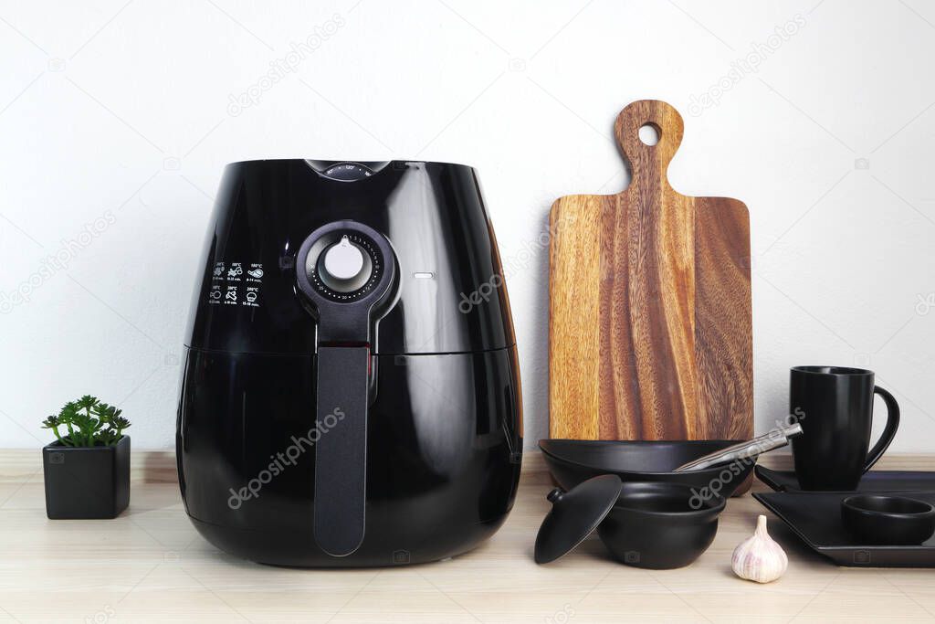 a black deep fryer or oil free fryer appliance, mug, dish and wooden tray are on the wooden table in the kitchen with a small plant in the pot ( air fryer ) with background of white cement wall