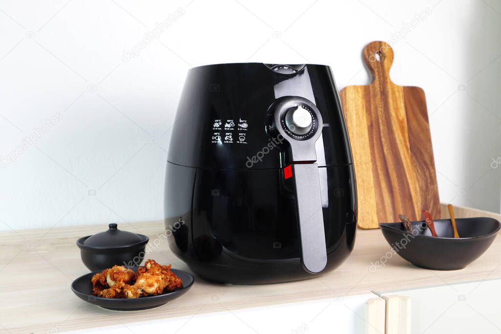 a black deep fryer or oil free fryer appliance, mug, dish and wooden tray are on the wooden table in the kitchen  with a dish of fried chicken wings on the background of white cement wall