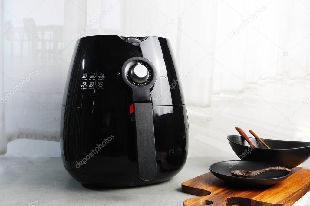 a black deep fryer or oil free fryer appliance, mug, dish and wooden tray are on the wooden table in the kitchen near background of white soft nice curtain and shadow of table and chairs dinning room 