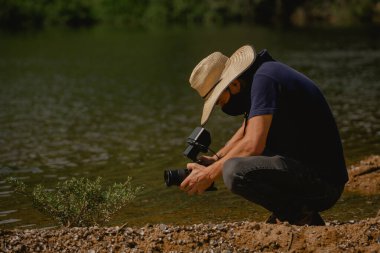 Güvenlik için rimel ve nehir kıyısında hasır şapka takan kameralı bir adam..