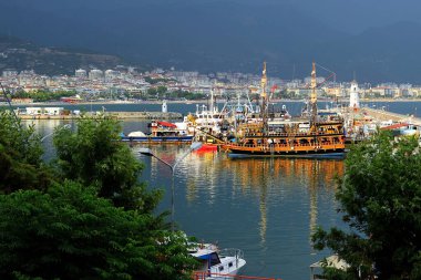 Güneş tarafından aydınlatılan Alanya limanındaki gemiler karanlık gök gürültüsünün arka planına karşı. Yeşil ağaçların önünde....