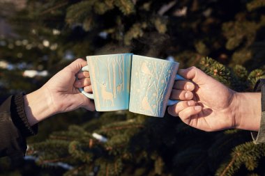 Kış ormanının arka planında kahve fincanları olan kadın ve erkeklerin elleri. Parkta karla kaplı ladin..
