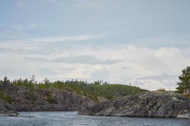 Suyun ortasında ağaçlarla kaplı taştan bir ada. Bulutlu mavi gökyüzü. Kuzey manzarası