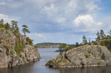 Suyun ortasında ağaçlarla kaplı taştan bir ada. Bulutlu mavi gökyüzü. Kuzey manzarası