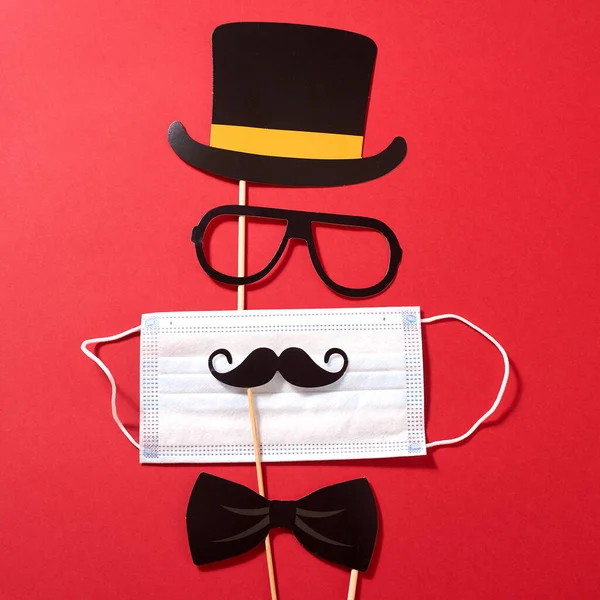 Paper hat, mustache, sunglasses and a bowtie with medical mask on the red background. Quarantine party.