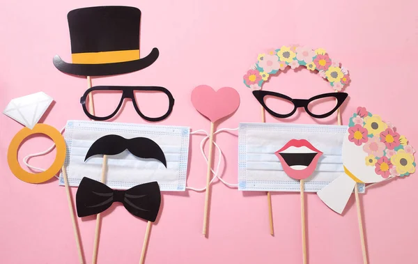 Paper groom and bride consisting of sunglasses, hat, mustache, lips with a medical mask, bridal bouquet, wedding ring on the pink background. Quarantine wedding party. Funny fashion.