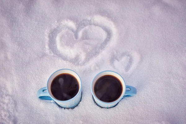 two blue mugs of steaming coffee in the snow with painted hearts in winter park.