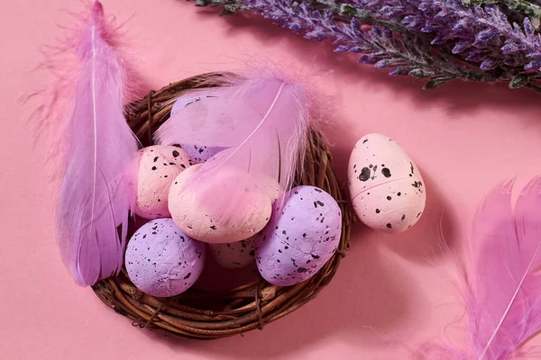 Close Up of Colorful Easter Eggs with pastel colored feathers in the nest with lavender bouquet on the pink background. Pastel colors, flat lay, top view.