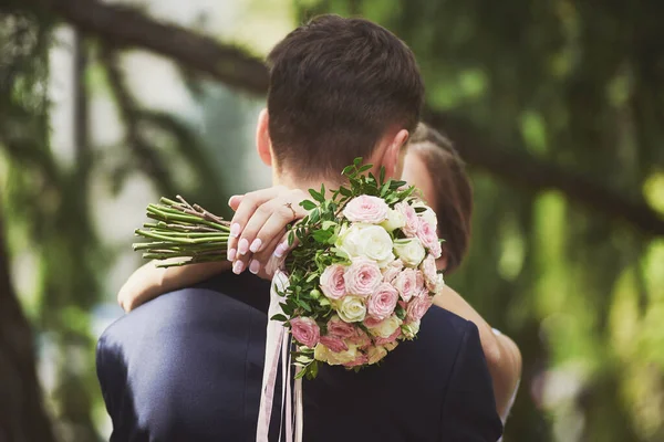 Gelin damadı kucaklıyor ve öpüyor. Gelinin elleri zarif bir buket beyaz ve pembe güllerle. Yüzüğe odaklan. Bulanık yeşil arkaplan.