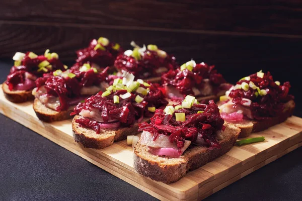 Traditional russian sandwiches with spicy beetroot salad, pickled onions and smoked herring. Bruschettas with beetroot and herring. Herring under a fur coat. Black background. Wooden cutting board.