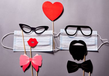 İki tıbbi maske, gözlüklü, dudaklı ve bıyıklı üzerinde kırmızı bir kalp olan gri bir arka plan.