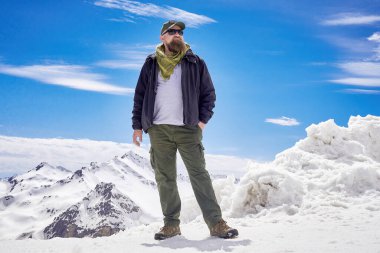 Turist kıyafeti giymiş sakallı bir adam, karla kaplı dağların arkasında poz veriyor. Seyahat ve macera modası