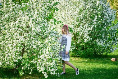 Beyaz kareli elbiseli ve pembe çelenkli güzel bir genç kız çiçek açan elma ağaçlarının arkasında poz veriyor. Bahar fotoğraf çekimi için bir fikir..