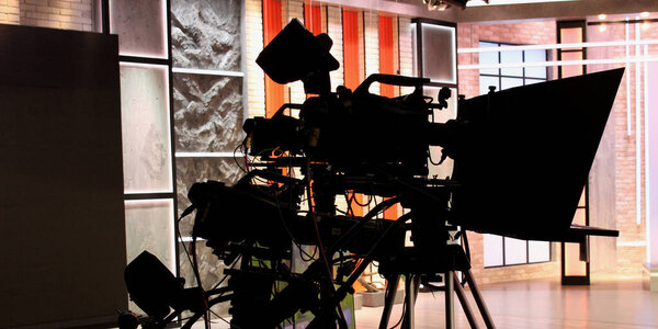 Silhouettes of studio cameras connected by wires to a broadcast console against the backdrop of brightly lit sets in a video studio
