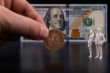 Erkek parmaklar, 100 dolarlık banknotun arka planına karşı sembolik insanların iki plastik figürüne 1 Amerikan doları para uzatıyor.