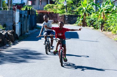 Mauritius Adası, 05.12.2015. İki genç çocuk, güneşli bir günde asfalt yolda bisikletlerinde gülümseyerek el kol geziyorlar..