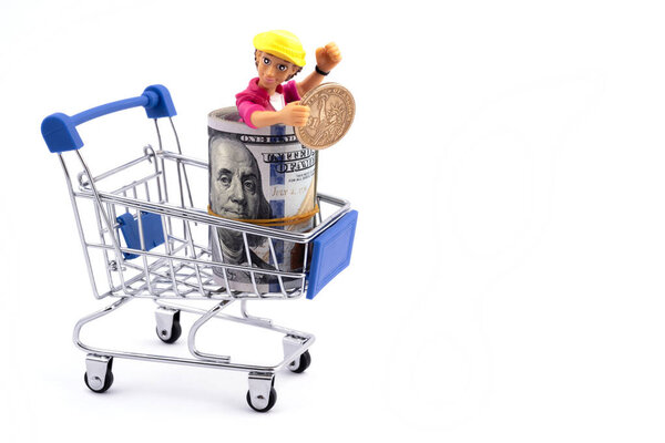 Символический пластиковый человек внутри пачки банкнот в 100 американских долларов с монетой в 1 доллар и миниатюрной корзиной для покупок на белом фоне