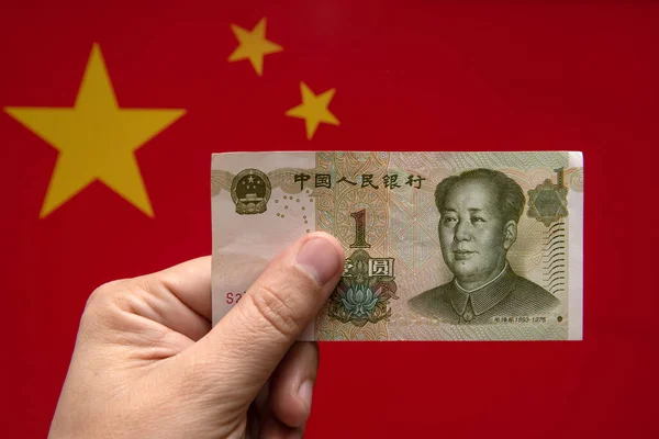 A man\'s hand holds a one yuan bill with a portrait of Mao against the background of a red Chinese flag