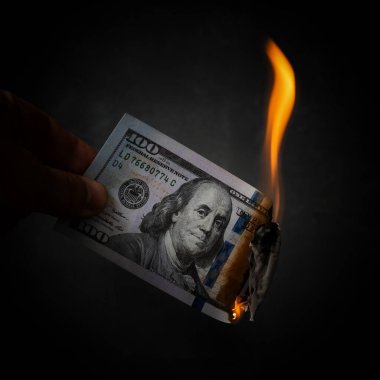Parmakların üzerinde Benjamin Franklin 'in siyah bir arkaplanda portresi olan 100 dolarlık yanan bir banknot var.